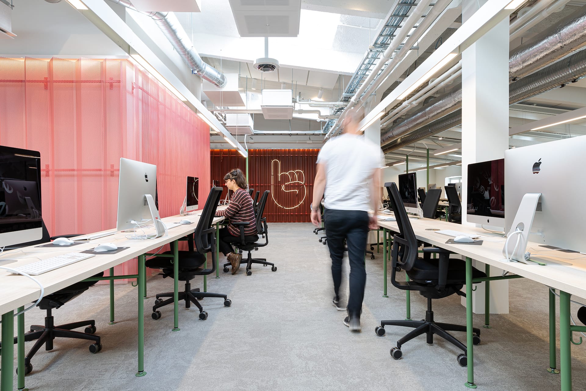 Interior view of Hive Helsinki, a modern open-plan office or coworking space. Rows of light-colored desks with green legs are equipped with Apple iMac computers. A person is seated and working at a desk on the left, while another person walks in the aisle towards the right. The space features exposed industrial ceilings, bright lighting, and a decorative neon hand sign on a pink textured wall.