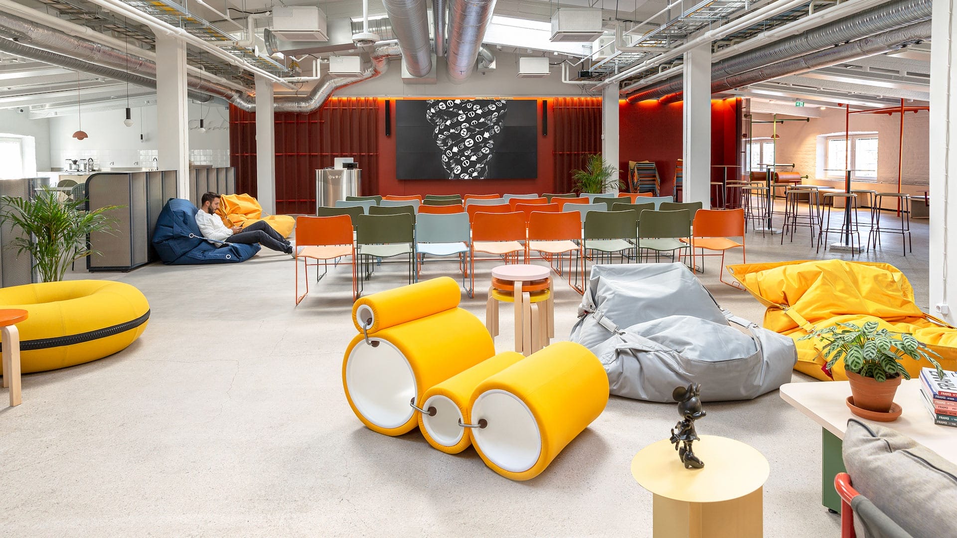 A spacious, modern lounge and auditorium area at Hive Helsinki, featuring a large screen at the back, rows of colorful chairs, and various casual seating options including bean bags and unique yellow tubular seating elements. Exposed ductwork and an industrial aesthetic are visible, with a person relaxing on a blue bean bag in the background.